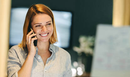 Fröhliche junge Geschäftsfrau, die fröhlich lächelt, während sie ein Telefongespräch führt. Erfolgreiche Geschäftsfrau, die mit ihren Geschäftspartnern in einem modernen Büro kommuniziert. - JLPSF10248