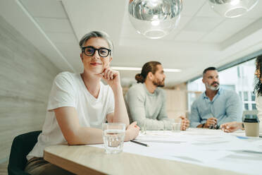 Female design professional looking at the camera while sitting in a meeting with her team. Group of creative businesspeople brainstorming while working on a new project. - JLPSF10253