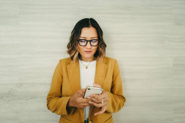 Ethnische Geschäftsfrau, die eine Textnachricht liest, während sie an einer Wand steht. Junge Unternehmerin, die ein Smartphone benutzt, während sie an einem modernen Arbeitsplatz arbeitet. - JLPSF10255