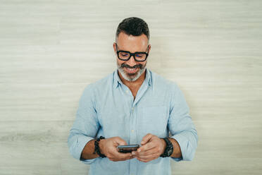 Reifer Geschäftsmann, der fröhlich lächelt, während er eine Textnachricht auf seinem Smartphone liest. Erfolgreicher Geschäftsmann, der mit seinen Mitarbeitern kommuniziert, während er an einem modernen Arbeitsplatz arbeitet. - JLPSF10259