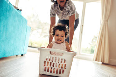 Niedliches Baby, das in einem Wäschekorb sitzt und von der Mutter geschoben wird. Nette Familie, die zusammen zu Hause spielt. - JLPSF10318