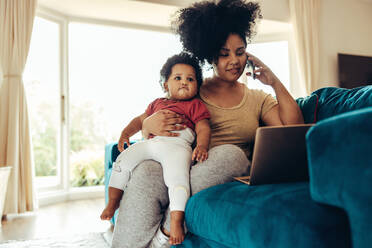 Eine afrikanische Mutter sitzt mit einem Baby auf dem Sofa, telefoniert und arbeitet am Laptop. - JLPSF10322