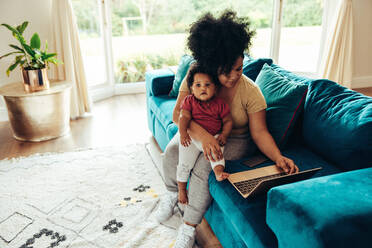 Frau mit Baby auf dem Sofa sitzend und Laptop benutzend, Mutter mit Baby bei der Arbeit zu Hause. - JLPSF10323