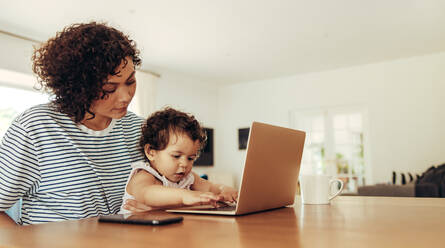 Mother watching baby pressing with laptop keyboard keys. Freelancer woman sitting with baby and laptop at home. - JLPSF10327