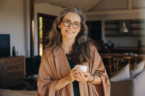 Portrait of a happy senior woman smiling at the camera while standing with a cup of tea in her hands. Cheerful senior woman enjoying a serene retirement at home. - JLPSF10332