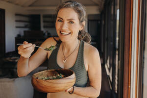 Glückliche Veganerin, die in die Kamera lächelt, während sie einen Gemüsesalat aus einer Schüssel isst. Ältere Frau, die nach einem Workout zu Hause ein pflanzliches Frühstück genießt. Ältere Frau, die ihren alternden Körper pflegt. - JLPSF10334