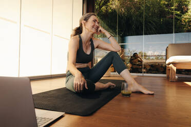 Glückliche ältere Frau, die lächelnd vor einem Laptop auf einer Übungsmatte sitzt. Ältere Frau, die eine Pause von einem Yogakurs zu Hause macht. Fröhliche ältere Frau, die einem Online-Fitness-Tutorial folgt. - JLPSF10338