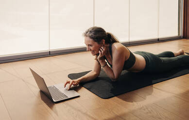 Elderly woman smiling while joining a virtual fitness class on a laptop. Happy senior woman following an online yoga tutorial at home. Cheerful mature woman lying on an exercise mat. - JLPSF10345