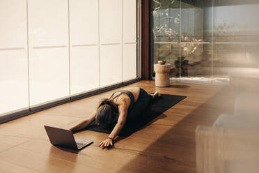 Woman doing a child's pose exercise during an online yoga class at home. Senior woman practicing a kneeling asana on a yoga mat. Mature woman following a virtual workout tutorial on a laptop. - JLPSF10348