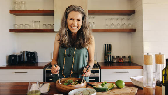 Excited senior woman smiling cheerfully while having some delicious vegan food. Mature woman serving herself a healthy buddha bowl at home. Woman taking care of her aging body with a plant-based diet. - JLPSF10362