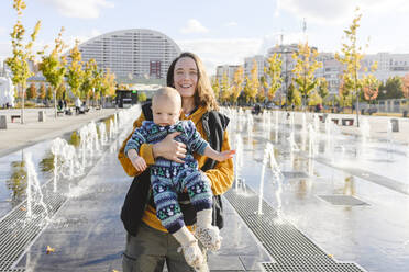 Happy mother and son enjoying with each other amidst fountains at urban park - EYAF02207