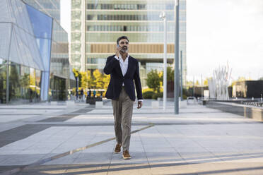 Mature businessman walking and talking on smart phone in front of office building - JCCMF07513