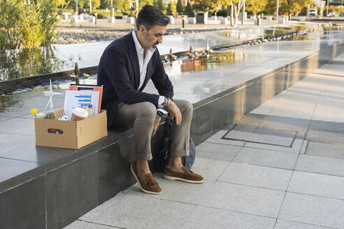 Trauriger Geschäftsmann mit Büromaterial in einer Kiste an der Wand sitzend - JCCMF07522
