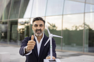 Smiling businessman showing thumbs up gesture holding wind turbine model - JCCMF07531
