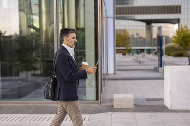 Businessman using smart phone walking in front of office building - JCCMF07533