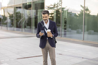 Businessman with disposable cup and mobile phone standing in front of office building - JCCMF07534