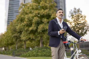 Smiling businessman with smart phone holding bicycle on footpath - JCCMF07541