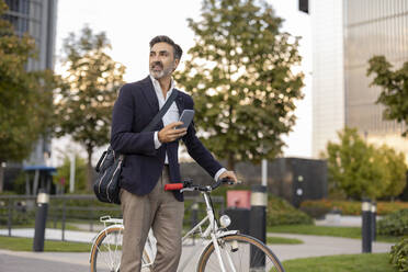 Mature commuter with smart phone and bicycle on footpath - JCCMF07542