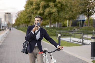 Businessman talking on speaker phone wheeling with bicycle on footpath - JCCMF07543