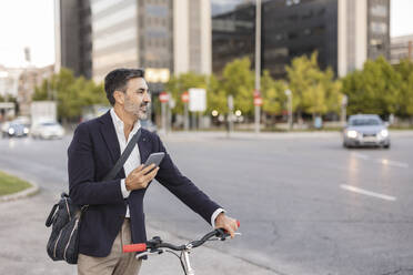 Smiling commuter with smart phone and bicycle at roadside - JCCMF07546