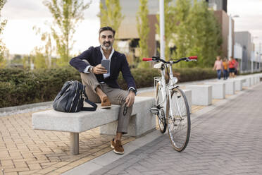 Lächelnder Geschäftsmann mit Smartphone auf einer Bank neben einem Fahrrad sitzend - JCCMF07548