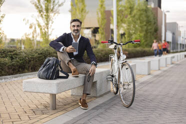 Smiling businessman with smart phone sitting on bench by bicycle - JCCMF07548