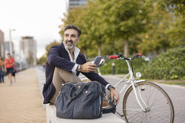 Happy businessman with smart phone resting on bench by bicycle - JCCMF07549