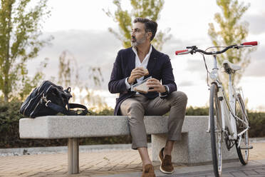 Businessman eating sandwich sitting on bench by bicycle - JCCMF07550