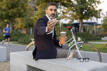 Geschäftsmann mit Einwegbecher und Sandwich auf einer Bank am Fahrrad - JCCMF07551