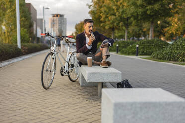 Geschäftsmann beim Mittagessen auf einer Bank sitzend neben einem Fahrrad auf einem Fußweg - JCCMF07552