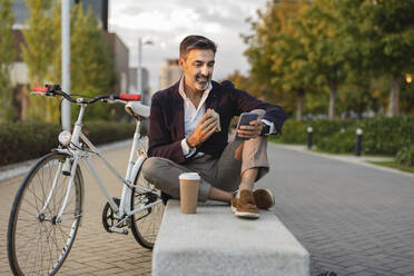 Lächelnder Geschäftsmann, der sein Smartphone benutzt und auf einer Bank mit dem Fahrrad zu Mittag isst - JCCMF07553