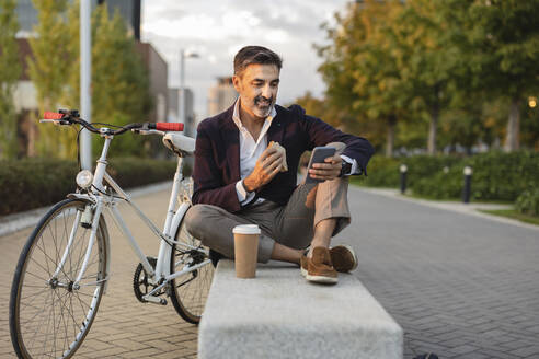 Lächelnder Geschäftsmann, der sein Smartphone benutzt und auf einer Bank mit dem Fahrrad zu Mittag isst - JCCMF07553