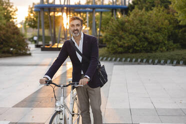 Smiling commuter wheeling with bicycle on footpath - JCCMF07556