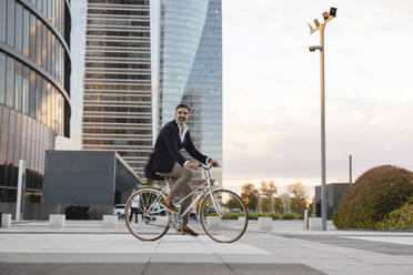 Mature commuter riding bicycle in front of office building at sunset - JCCMF07561