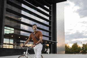 Nachdenklicher Geschäftsmann, der mit dem Fahrrad vor einem Gebäude radelt - JCCMF07567