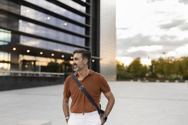 Smiling businessman with hands in pockets standing by office building - JCCMF07568