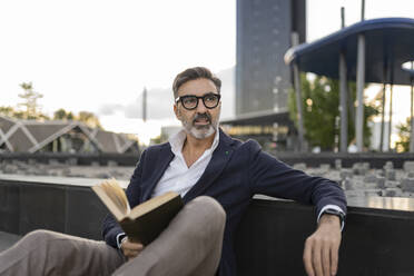 Thoughtful businessman with book by wall - JCCMF07580