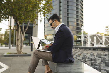 Businessman using laptop sitting on wall - JCCMF07584