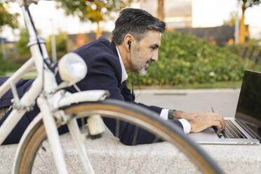 Businessman with headphones using laptop lying on bench by bicycle - JCCMF07595