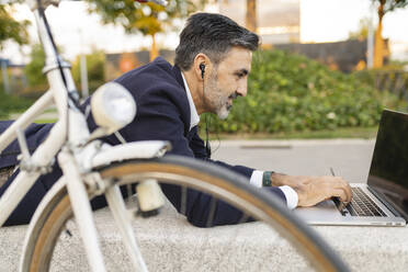 Geschäftsmann mit Kopfhörern und Laptop auf einer Bank neben einem Fahrrad liegend - JCCMF07595
