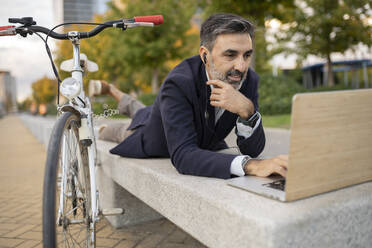 Businessman working on laptop by bicycle near bench - JCCMF07597