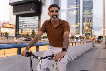 Smiling businessman sitting on bicycle at footpath - JCCMF07603