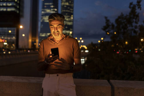 Smiling businessman with mobile phone in city at night - JCCMF07610