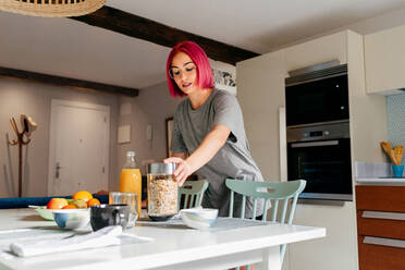 Junge Frau mit hellrosa Haaren, die ein Glas mit Getreide vom Tisch nimmt, während sie das Frühstück in einer modernen Wohnung zubereitet - ADSF39408