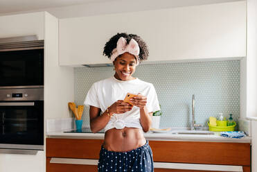 Glückliche afroamerikanische Frau, die lächelt und auf ihrem Smartphone surft, während sie in der Küche einer modernen Wohnung steht - ADSF39409