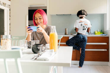 Erfreute junge Frau mit leuchtend rosa Haaren, die lächelt und auf ihrem Smartphone surft, während sie am Tisch in der Nähe eines kochenden afroamerikanischen Freundes beim Frühstück zu Hause sitzt - ADSF39410