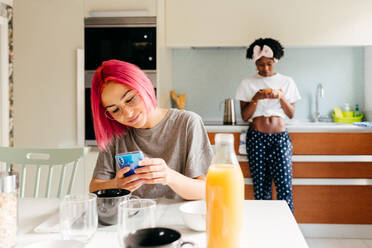 Delighted young lady with bright pink hair smiling and browsing smartphone while sitting at table near cooking African American friend during breakfast at home - ADSF39411