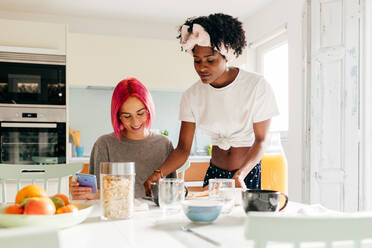 Erfreute junge Frau mit leuchtend rosa Haaren, die lächelt und auf ihrem Smartphone surft, während sie am Tisch in der Nähe eines kochenden afroamerikanischen Freundes beim Frühstück zu Hause sitzt - ADSF39413