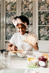 Happy African American lady smiling and adding fresh oatmeal in bowl while sitting at table and having breakfast at home - ADSF39415
