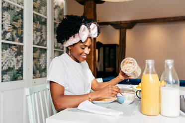 Glückliche afroamerikanische Frau, die lächelt und frische Haferflocken in eine Schüssel gibt, während sie am Tisch sitzt und zu Hause frühstückt - ADSF39416